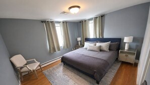 Ein Schlafzimmer mit einem Bett, Nachttischen, einem Sessel und einem Fenster mit Vorhängen.