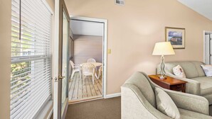 A room with a sliding glass door leading to a balcony, a sofa, a lamp, and a framed picture on the wall. - Luxury Condo with Golf Course Views, Pools & Resort Access! (New Bern)