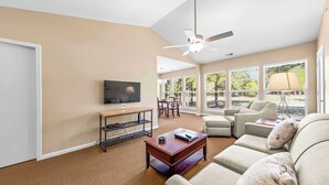 A living room with a sofa, coffee table, TV, and a ceiling fan. - Luxury Condo with Golf Course Views, Pools & Resort Access! (New Bern)