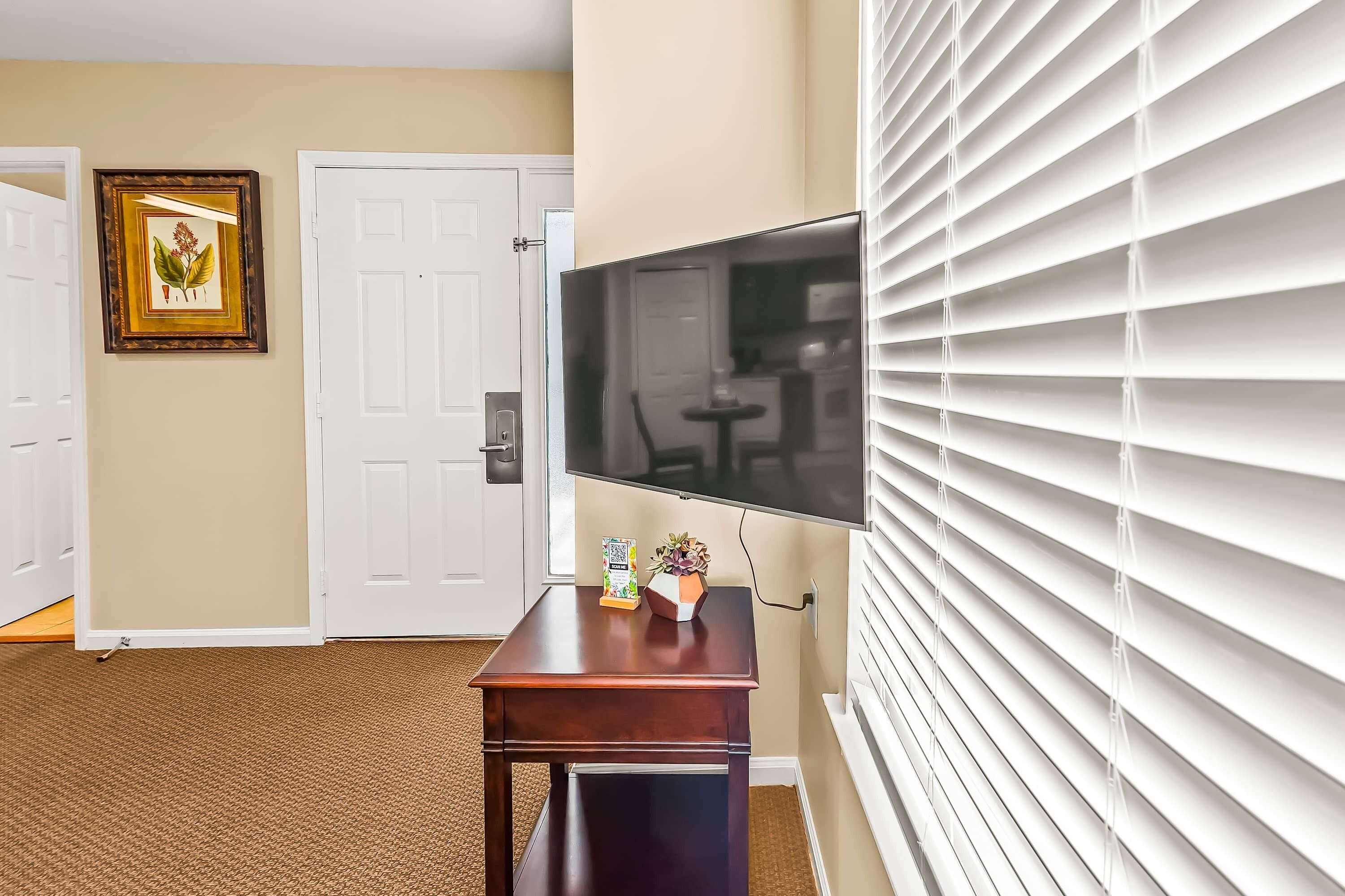 A room with a wooden table, a framed botanical print, a door, and a wall-mounted TV.