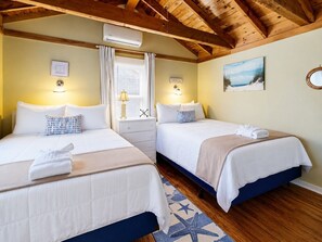 A hotel room with two beds, a wooden ceiling, an air conditioning unit, a bedside table with a lamp, and a beach-themed wall decoration. - Coastal Shorelands Retreat + Walk-to-Beach Access and Pool (Kennebunk)
