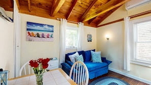 A cozy room with a wooden ceiling, a blue sofa with patterned cushions, a dining table with a vase of red roses, and a beach-themed painting on the wall. - Coastal Shorelands Retreat + Walk-to-Beach Access and Pool (Kennebunk)