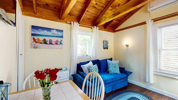 Ein gemütlicher Raum mit Holzbalkendecke, einem blauen Sofa mit gemusterten Kissen, einem Esstisch mit einer Vase roter Rosen und einem Strandbild an der Wand.