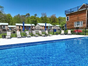 Ein Schwimmbecken mit Liegestühlen und Sonnenschirmen, Holzbauten und ein eingezäuntes Areal.