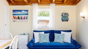 A room with a blue sofa, a white chair, a small table, a painting of beach chairs, a window with curtains, and a wall sign saying 'The Beach is my Happy Place.' - Coastal Shorelands Retreat + Walk-to-Beach Access and Pool (Kennebunk)