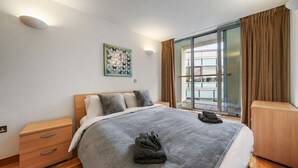 Ett hotellrum med en säng, ett träbord vid sängen, en väggmonterad lampa, ett fönster med gardiner och utsikt över omgivningen.
