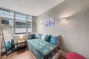 Un dormitorio con cama, escritorio, silla, ventana y un cuadro en la pared.
