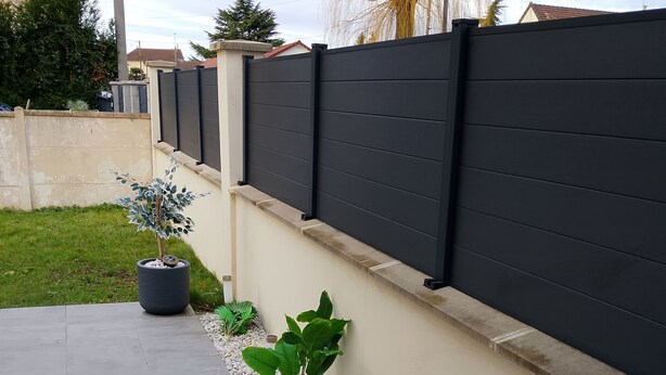 Un espace extérieur aménagé avec un revêtement pavé, un petit muret, des plantes en pot et un poteau métallique.