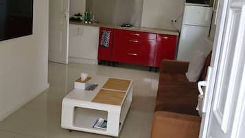 A modern living room with a white coffee table, a brown sofa, and a kitchen area with red cabinets and white appliances.