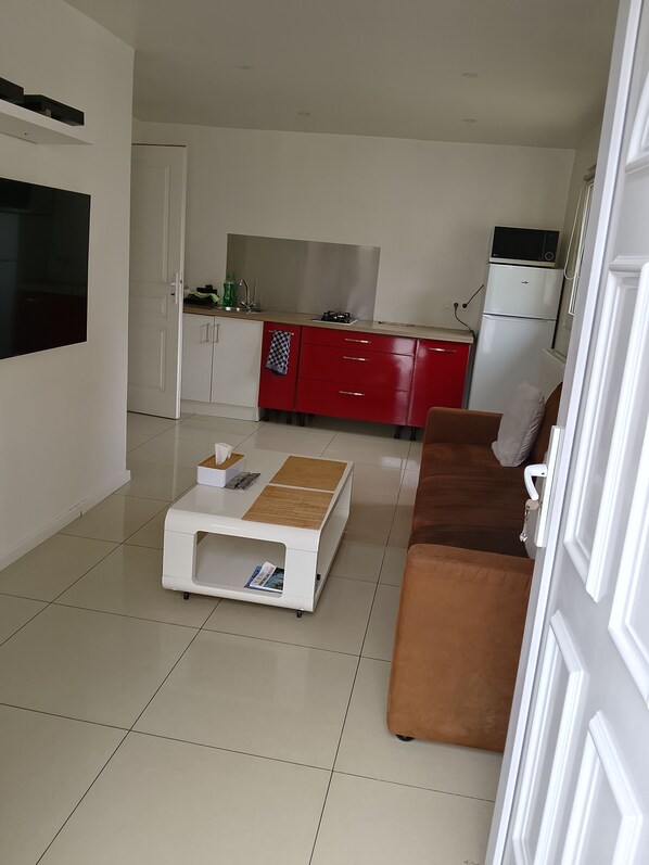 A modern living room with a white coffee table, a brown sofa, and a kitchen area with red cabinets and white appliances.