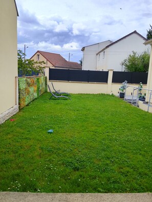 Un jardin à l’arrière d’une maison, avec une pelouse, une clôture noire et une maison dont le toit est rouge.