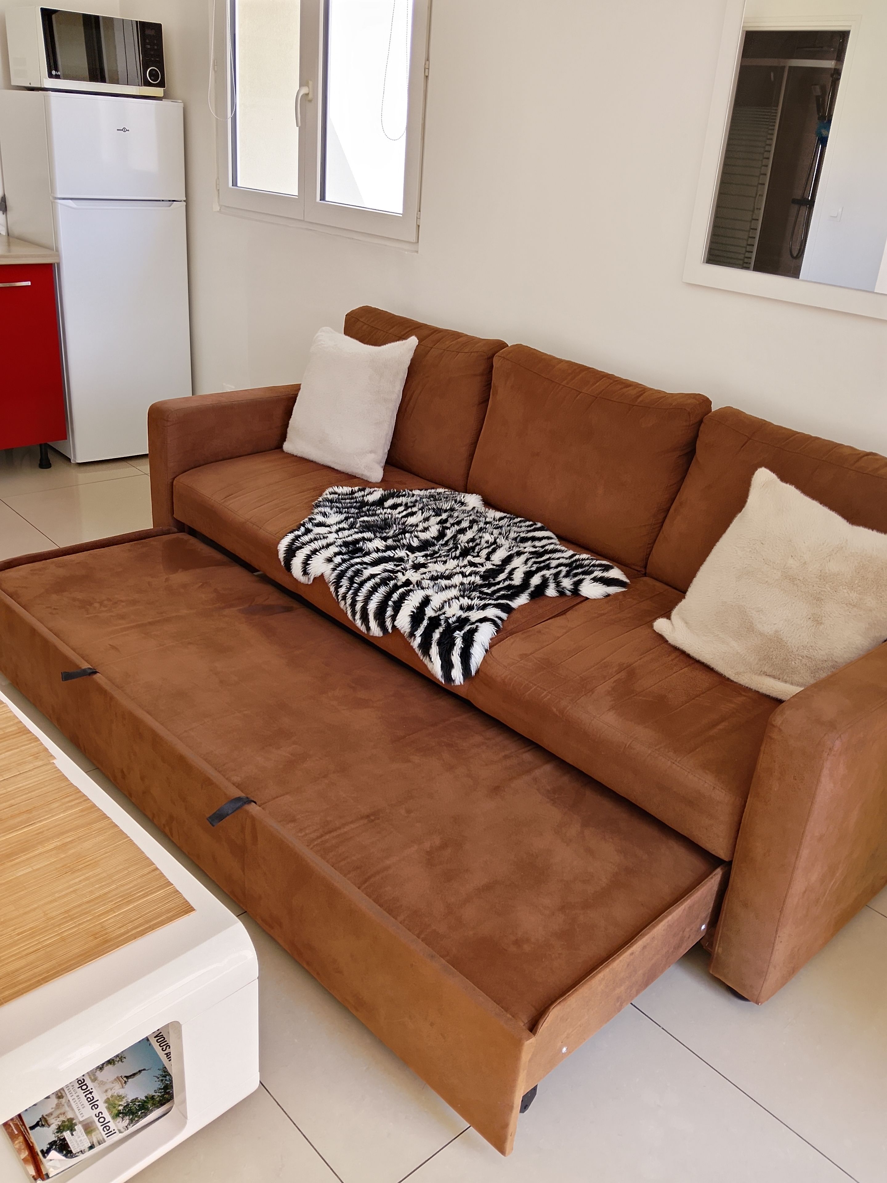 A brown sofa with a zebra-patterned throw blanket and two pillows, a white cabinet, and a microwave on top of a white refrigerator.