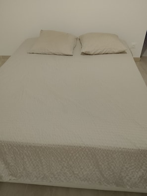 Un lit simple avec un matelas blanc et deux oreillers, placé contre un mur uni, avec une prise électrique visible.