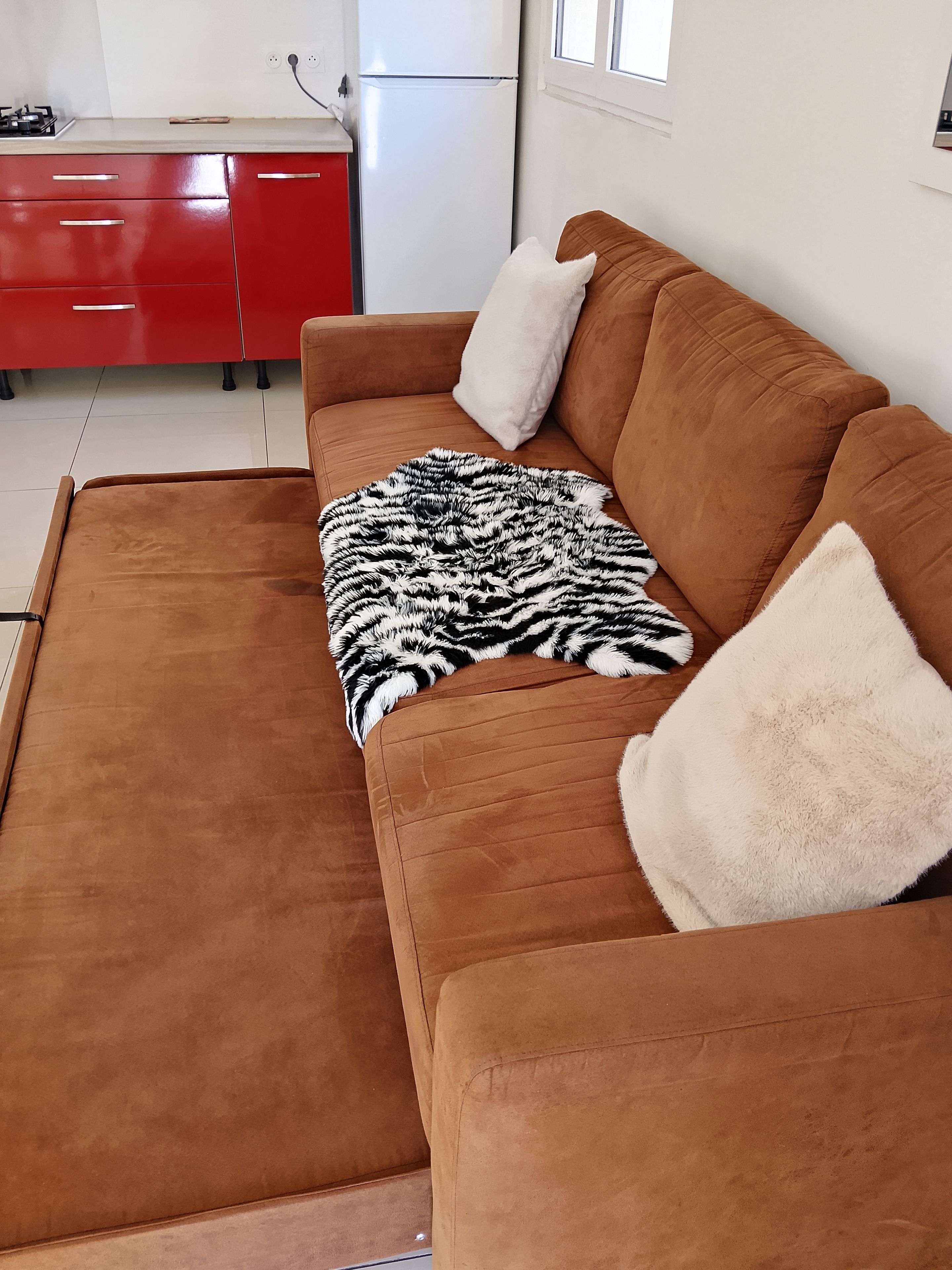 Een bruine bank met een zwart-wit deken en twee kussens, een rode kast en een witte koelkast in een kamer met witte muren en een raam.