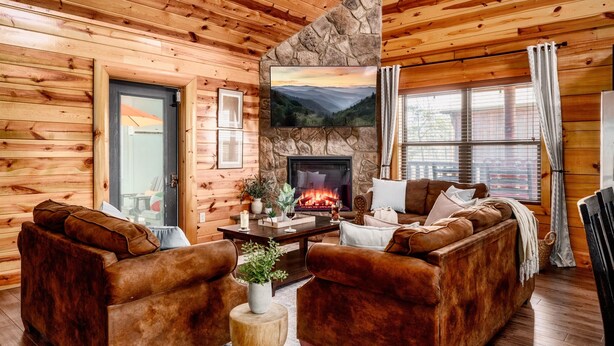 A cozy log cabin interior with a fireplace, wooden walls, and a stone accent wall. - Indoor Pool + Arcade Loft | 4 En-Suite BRs | Gated Bear Crossing Cabin (Sevierville)