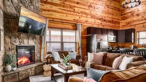 A cozy living room with a stone fireplace, wooden ceiling, and a kitchen area with modern appliances. - Indoor Pool + Arcade Loft | 4 En-Suite BRs | Gated Bear Crossing Cabin (Sevierville)