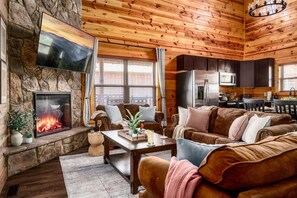 Ein gemütliches Wohnzimmer mit Kamin aus Stein, Holzboden und einer Küchenzeile mit modernen Geräten.