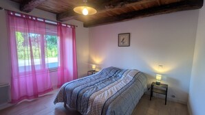 Un dormitorio con una cama, dos lámparas de noche, un ventanal con cortinas rosadas, techo de madera y un cuadro en la pared.