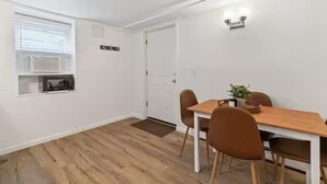 A room with a wooden floor, white walls, a dining table with chairs, a small window with blinds, and a door.