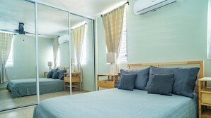 Een slaapkamer met een groot bed, een kleiner bed, een nachtkastje, een lamp en een plafondventilator.