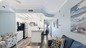 Una cocina moderna con armarios blancos, una isla central y una zona de comedor con sofá gris y cojines estampados.