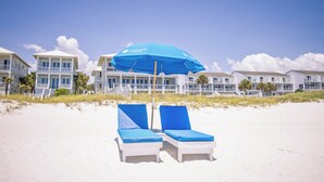 Una playa con dos sillas de playa blancas bajo un paraguas azul, edificios blancos con balcones y un cielo despejado.