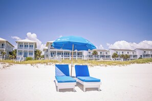 A beach with two white lounge chairs under a blue umbrella, white buildings with balconies, and a clear sky. - Carillon Inn 515. The Flagship Unit at the Inn. Corner Unit. 5th floor. Specials (Panama City Beach)