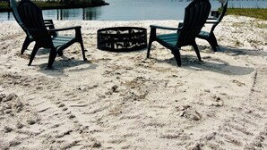 Uma praia com duas cadeiras Adirondack voltadas para águas calmas, uma faixa de areia e uma linha de árvores ao longe.