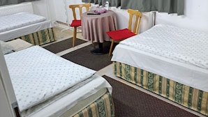 A hotel room with two single beds, a round table with a red tablecloth, two wooden chairs, a radiator, and a window with curtains.