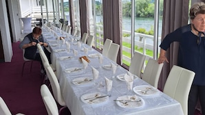 A long dining table with chairs, set for a meal, in a room with large windows overlooking a body of water.