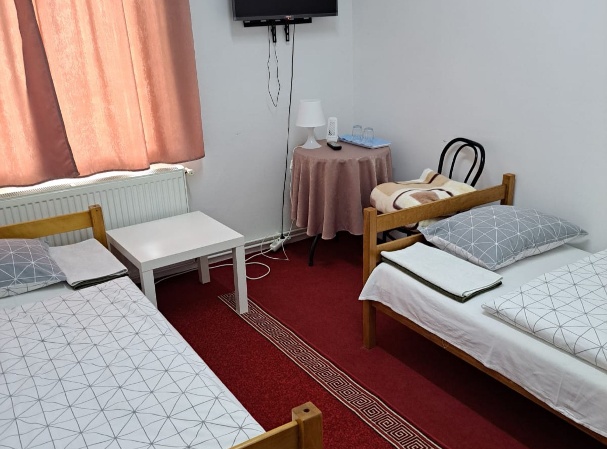 A room with two single beds, a small table with a lamp, a TV mounted on the wall, and a radiator.