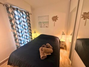 A bedroom with a bed, a nightstand, a lamp, a wall-mounted picture, and a patterned curtain. - Asteria by Pom, pkg, clim, wifi (Agde)