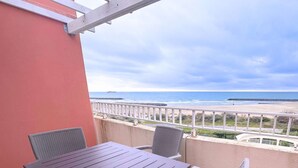 A balcony with a table and chairs, overlooking a beach with a rocky breakwater and a ship in the distance. - Asteria by Pom, pkg, clim, wifi (Agde)