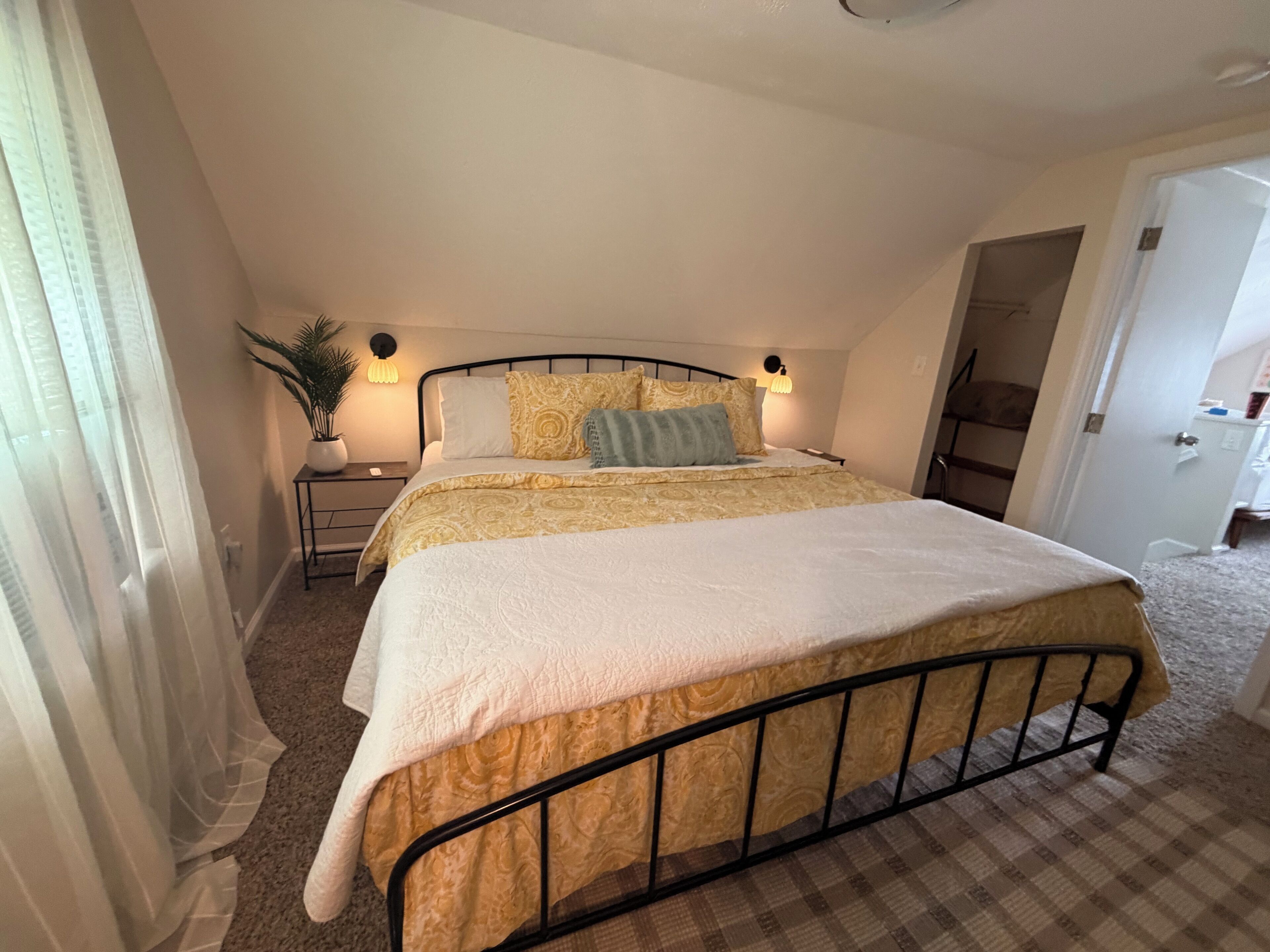 Une chambre à coucher avec un lit, des tables de chevet, une plante en pot et une porte donnant sur une autre pièce.