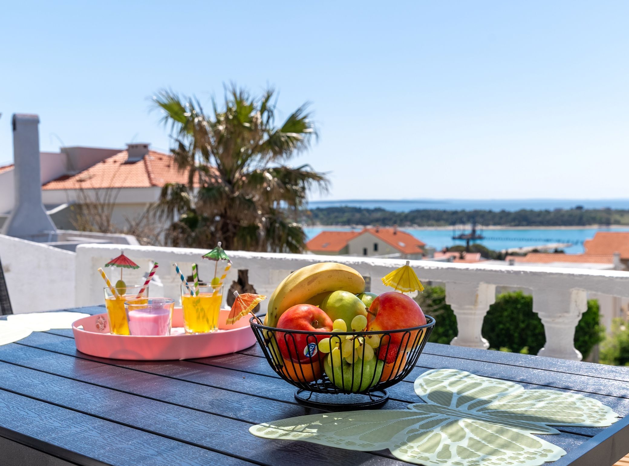 A table with a fruit bowl and drinks, overlooking a coastal view with buildings and palm trees.