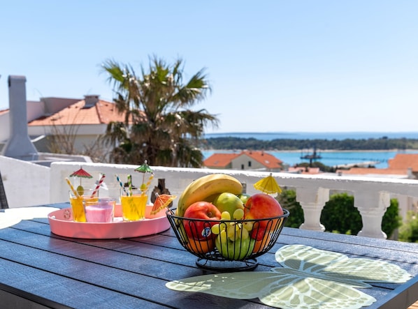 A table with a fruit bowl and drinks, overlooking a coastal view with buildings and palm trees.