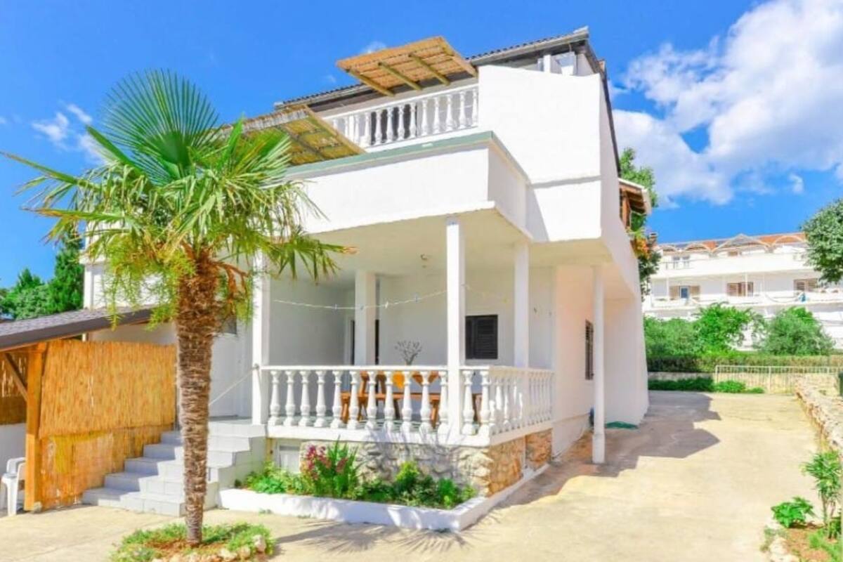 A two-story building with a balcony, a palm tree, and a stone wall.