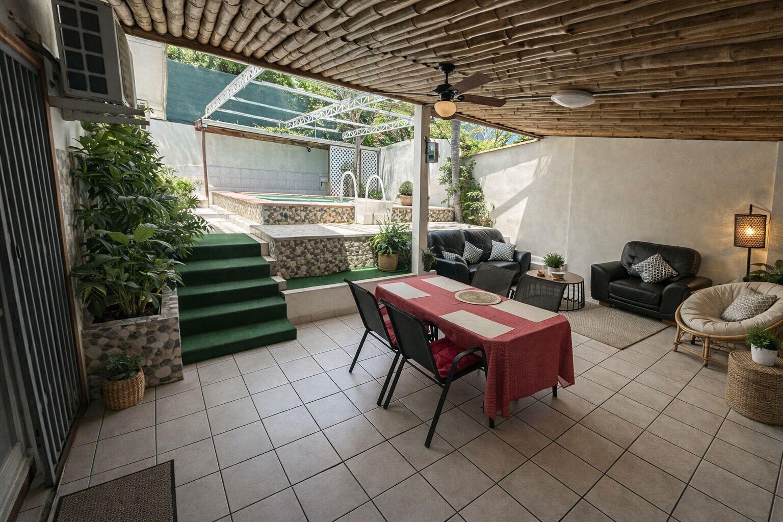 Un patio couvert comprenant une table à manger, des chaises, un canapé et un fauteuil. Des marches mènent à une zone abritant une piscine, et des plantes en pot sont disposées dans cet espace.