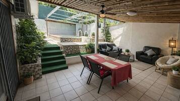 Un patio couvert comprenant une table à manger, des chaises, un canapé et un fauteuil. Des marches mènent à une zone abritant une piscine, et des plantes en pot sont disposées dans cet espace.
