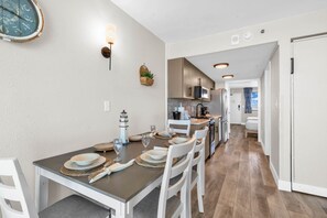 A modern kitchen with white cabinets, a dining area with a table set for four, and a hallway leading to a bedroom. - Compass Cove 1466: Beach View (Myrtle Beach)