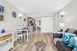 A modern living room with a brown leather sofa, a dining area with white chairs, a kitchen in the background, and a clock on the wall. - Compass Cove 1466: Beach View (Myrtle Beach)