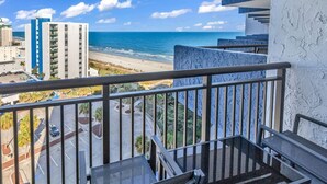 Un balcon avec vue sur la plage et l’océan, un parking et un immeuble à la façade bleue et blanche.