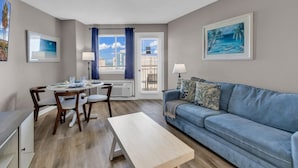 Un salon avec un canapé bleu, une table à manger avec des chaises, une table basse et une vue sur l’extérieur à travers une fenêtre avec des rideaux.