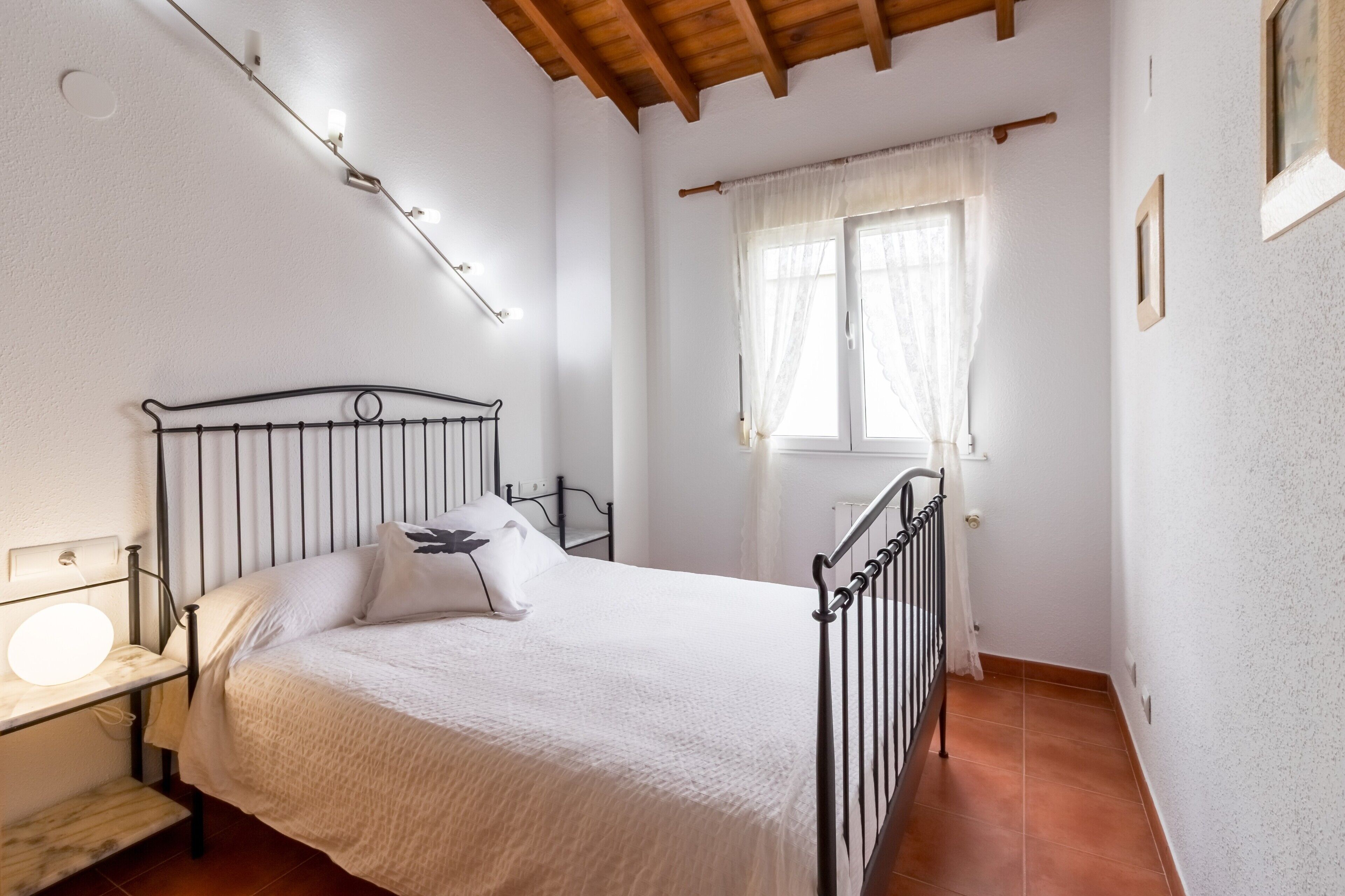 A bedroom with a bed, bedside lamp, window with curtains, and a wooden ceiling.