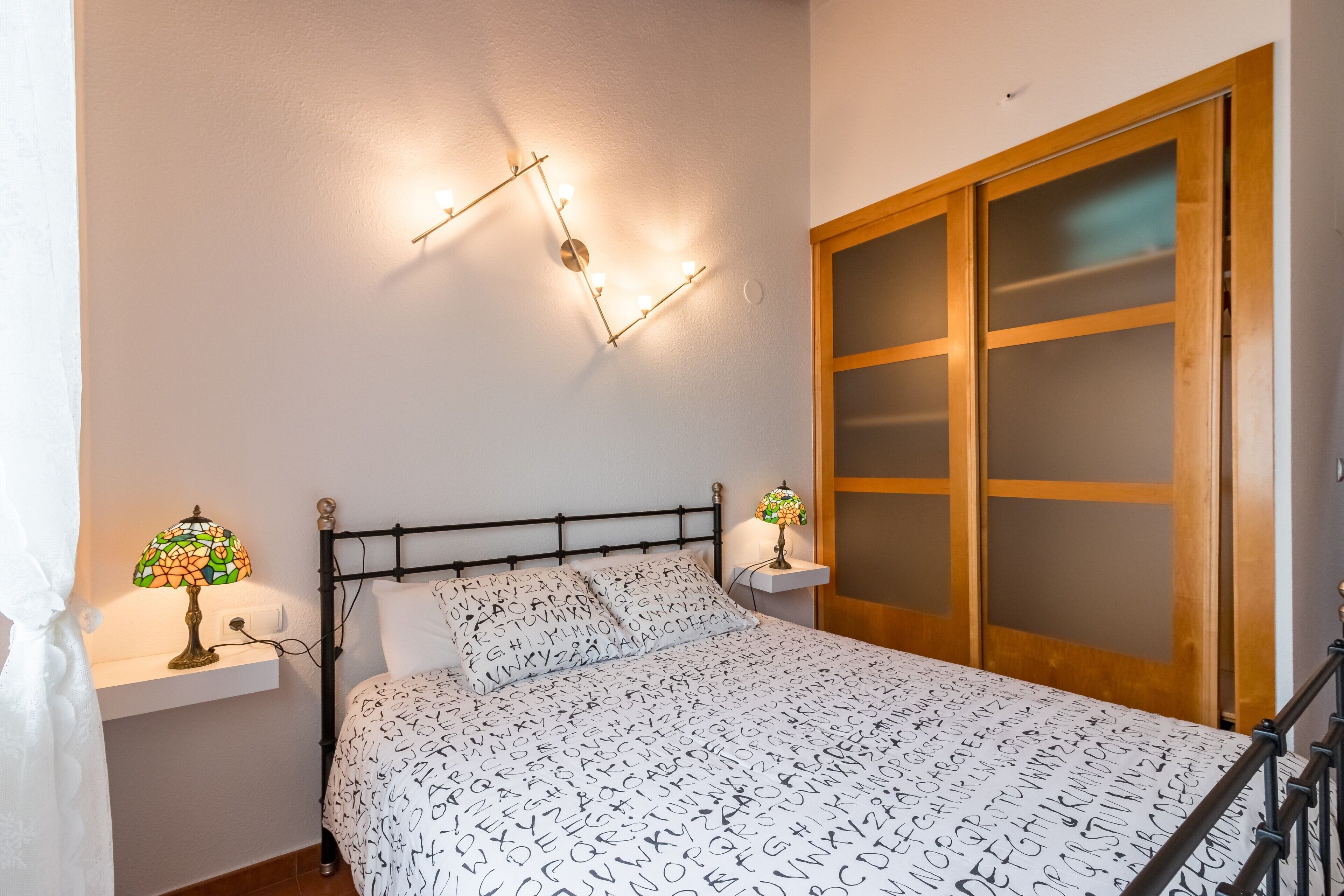 Un dormitorio con cama, dos lámparas de noche, una lámpara de pared y un armario de madera con puertas de vidrio esmerilado.