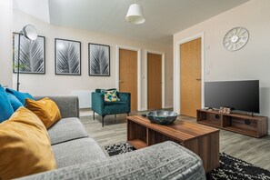 Ein modernes Wohnzimmer mit grauem Sofa, einem türkisfarbenen Sessel, einem Holztisch, einem Flachbildfernseher auf einem Holzgestell und einem großen Fenster mit Blick ins Freie.