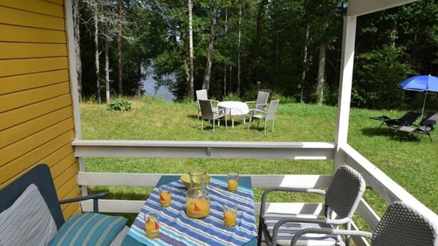 A covered outdoor patio with a table set for two, chairs, and a view of a grassy area with a table and chairs in the background. - Vacation home on the lake with rowing boat! (Nye)