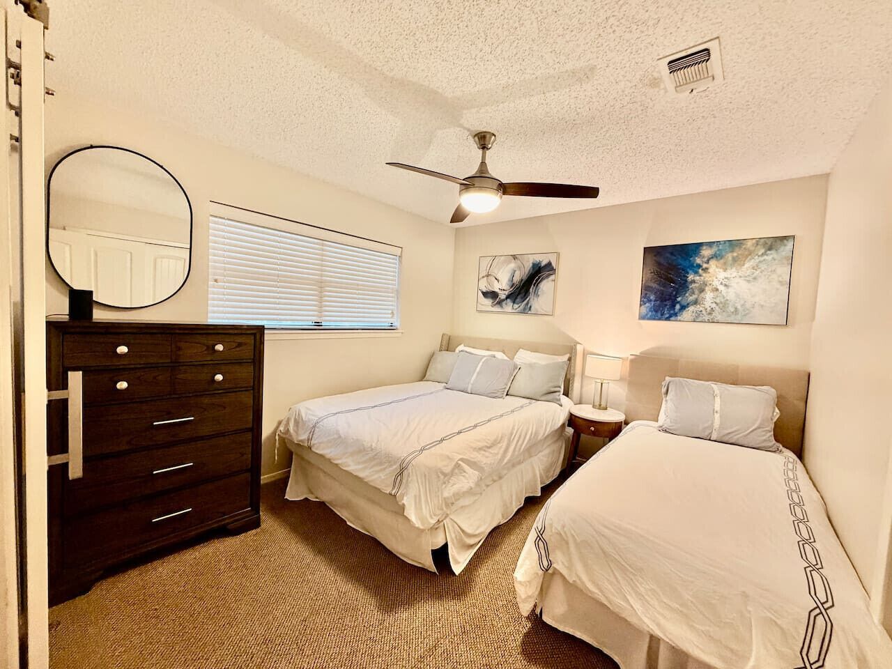 A bedroom with two beds, a dresser, a ceiling fan, and artwork on the walls.