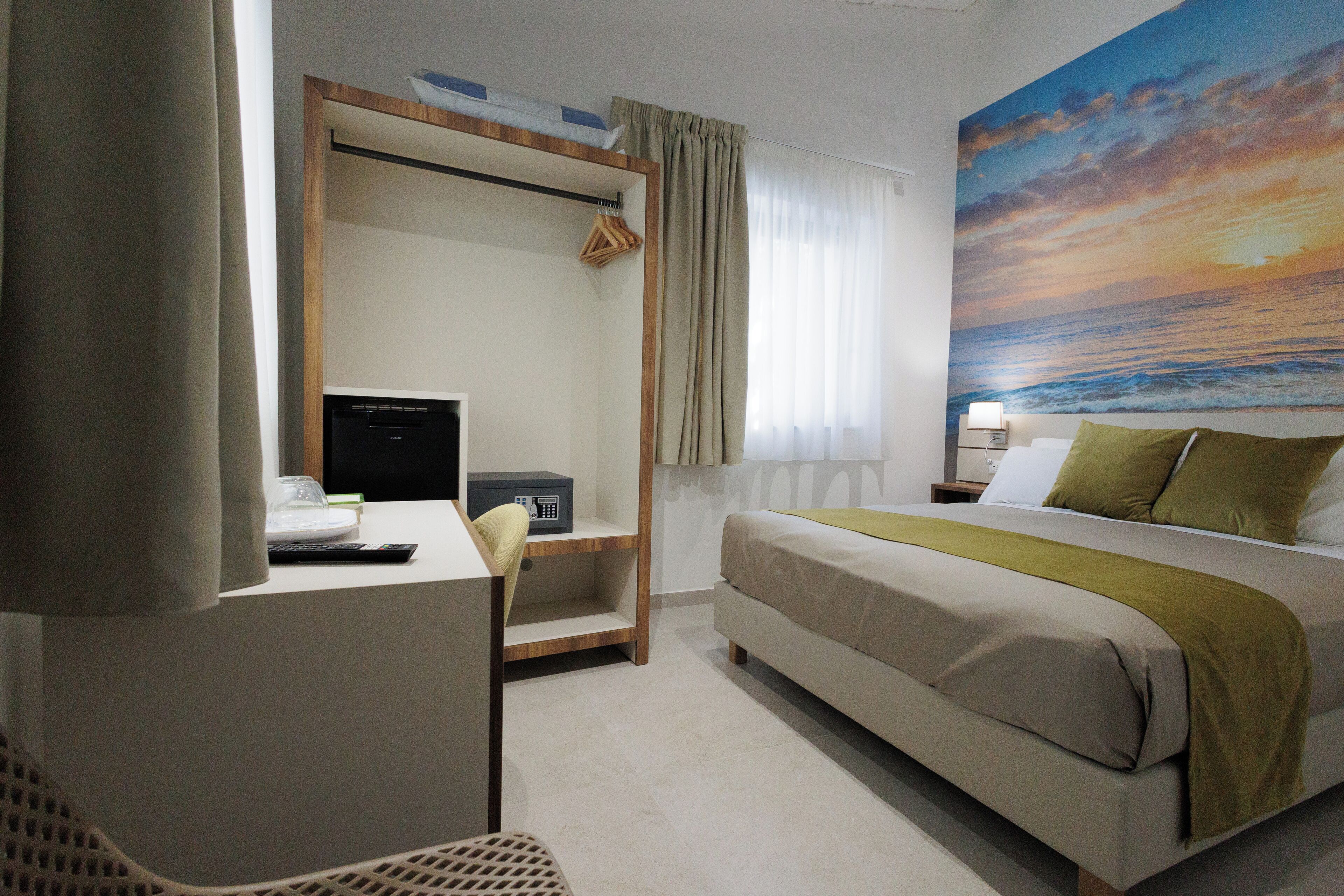 Une chambre d’hôtel avec un lit, un bureau, une chaise et un tableau représentant un coucher de soleil sur l’océan.