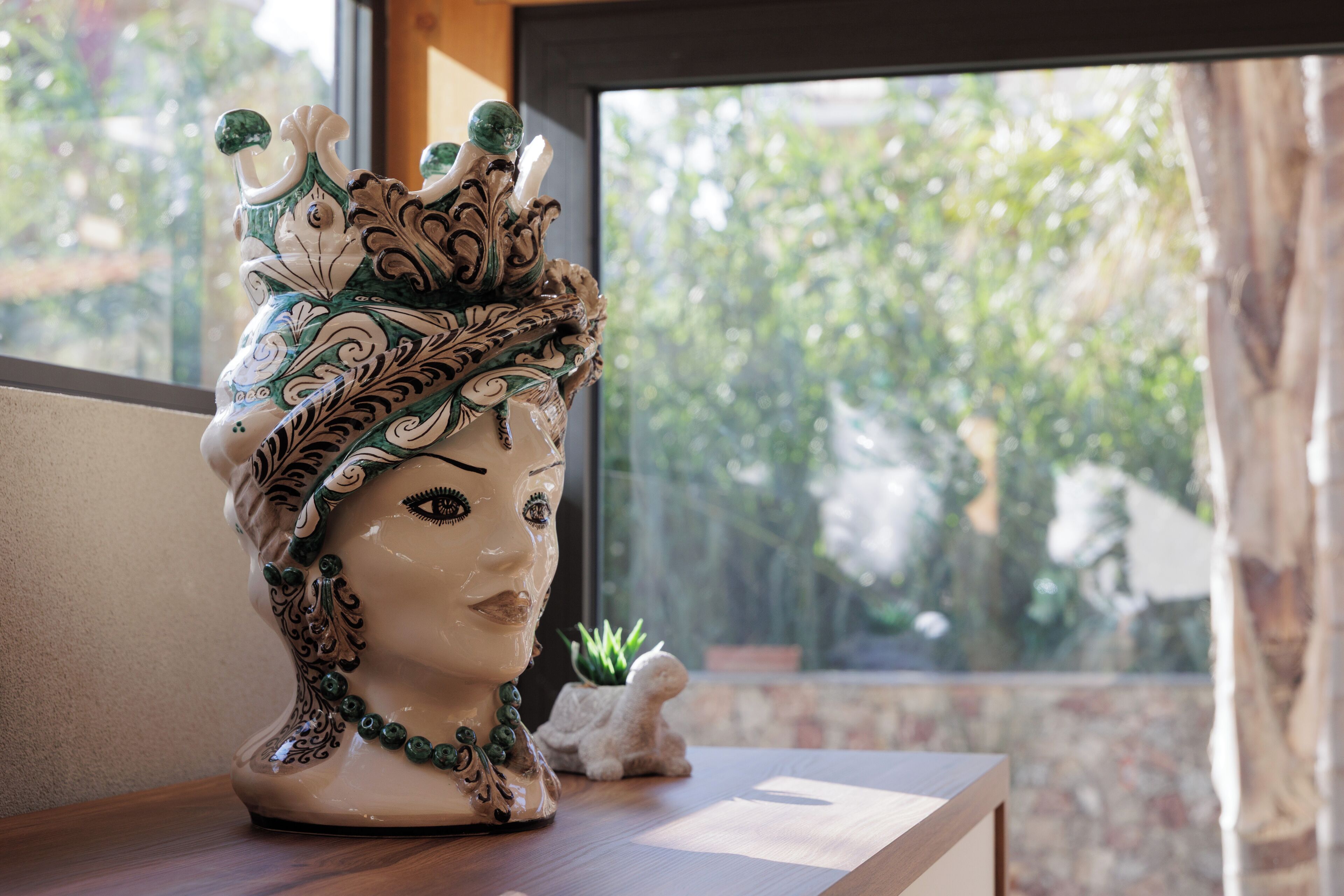 Busto in ceramica con decorazioni elaborate, posizionato su un piano di legno vicino a una finestra che si affaccia sul verde.